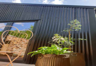 Capanna in legno nero con finestra riflettente, pianta in vaso e sedia in metallo a Blackbarn Wood, Regno Unito.