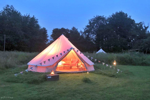 A cozy teepee tent glows with warm light at dusk on a grassy field, decorated with bunting and lanterns.