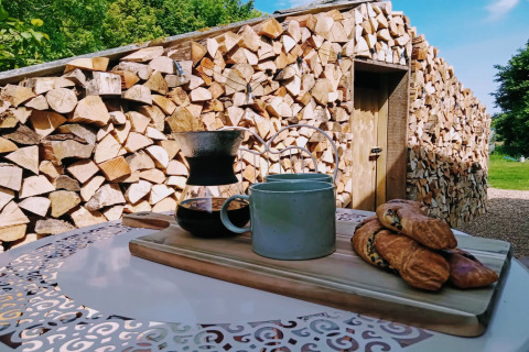 Colazione all'aperto presso Blackbarn Wood Lodge in UK con caffè, tazza e dolci davanti a legna accatastata.