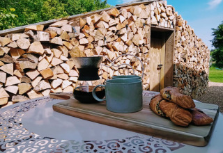Colazione all'aperto presso Blackbarn Wood Lodge in UK con caffè, tazza e dolci davanti a legna accatastata.