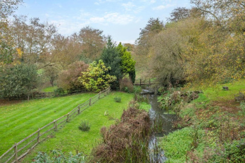Foto de un campo verde con un arroyo, árboles y un puente de madera en un entorno campestre tranquilo.
