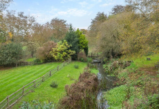 Photo of a grassy field with a stream, trees, and wooden bridge in a peaceful countryside setting.