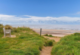 View from a grassy path with bench leading down to a sandy beach under a blue sky with clouds.