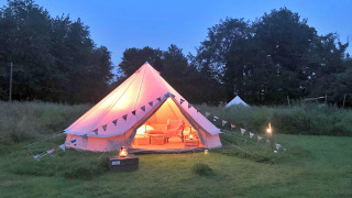 A glowing teepee tent in a grassy field at dusk, decorated with bunting and cozy furnishings inside.