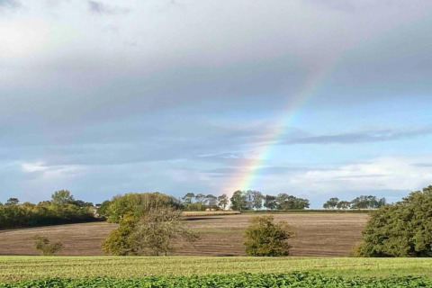 View of fields with a rainbow near Bulrush Bell Tent, a tipi tent in the UK, under a cloudy blue sky.