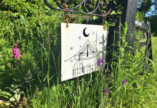 A sign featuring a tipi tent illustration hangs on a gate, surrounded by tall grass and wildflowers.