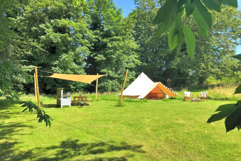 A cozy teepee tent on a grassy field with picnic table, sunshade, and chairs, surrounded by tall trees.