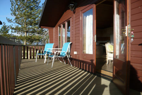 Terrazza soleggiata di una baita in legno con sedie blu, tavolo e porte aperte verso l’interno accogliente.