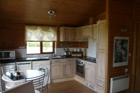 Cucina di baita in legno con mobili chiari, elettrodomestici moderni, tavolo tondo e tenda floreale alla finestra.