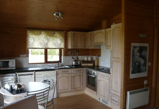 Cucina di baita in legno con mobili chiari, elettrodomestici moderni, tavolo tondo e tenda floreale alla finestra.
