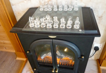 A glass chessboard with clear pieces sits atop a black stove in a wood-paneled cabin.