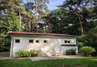 Uno chalet bianco chiamato Het Beerenbos in Belgio, circondato da alberi alti e sedie da giardino.