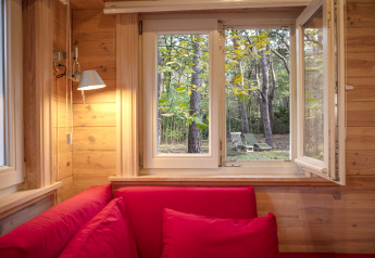 Zicht vanuit een houten chalet met rode zetels op het bos en tuinmeubelen door een open raam.