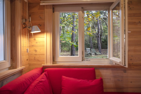Vista da uno chalet in legno con divani rossi attraverso una finestra aperta sul bosco e sedie da giardino.