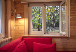 Vista da uno chalet in legno con divani rossi attraverso una finestra aperta sul bosco e sedie da giardino.