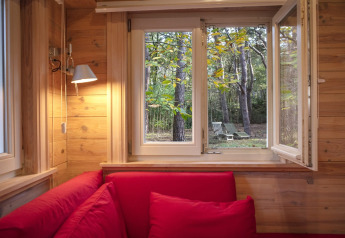 Vista desde un chalet de madera con sofás rojos, mirando por una ventana abierta hacia el bosque y sillas.