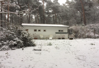 Chalet Het Beerenbos in Belgio innevato, con alberi, piante e sedie da giardino bianche all’esterno.