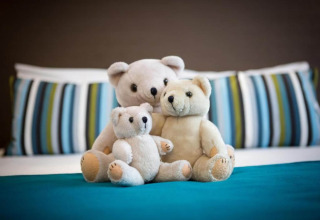 Tre orsetti di peluche seduti insieme su un letto con cuscini a righe nello chalet Het Beerenbos in Belgio.
