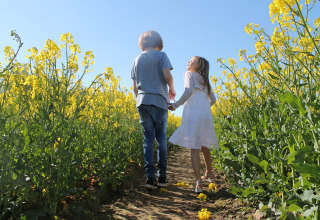 Twee kinderen wandelen hand in hand tussen gele bloemen bij Platz im Grünen 2 in Duitsland onder een blauwe hemel.