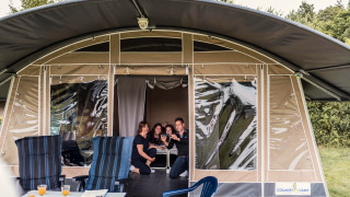 Una familia sonríe y se toma una selfie en la entrada de una tienda safari con sillas y mesa afuera.