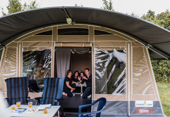 Une famille sourit et prend un selfie à l'entrée d'une tente safari, avec chaises et table à l'extérieur.