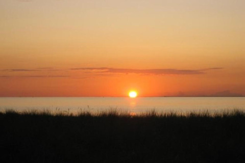 Puesta de sol vista desde una cabaña, con silueta de hierba oscura y agua tranquila bajo el cielo anaranjado.