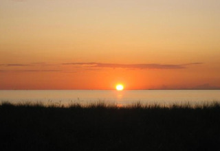 Puesta de sol vista desde una cabaña, con silueta de hierba oscura y agua tranquila bajo el cielo anaranjado.