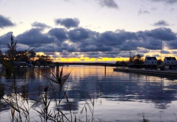 Coucher de soleil sur un site de glamping avec lodges près de l'eau et roseaux au premier plan.