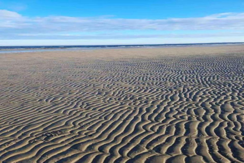 Strandfoto met geribbelde zandpatronen en blauwe lucht, genomen bij een glamping-accommodatie aan zee.