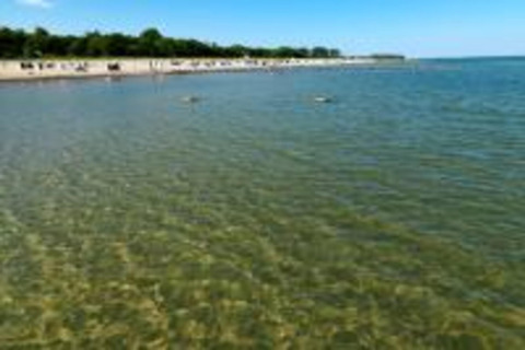 Clear, shallow water at a beach near a cabin with people on the shore and green trees in the background.
