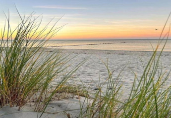Solnedgang over stranden med marehalm, roligt hav og blød sand ved et glampingsted nær kysten.