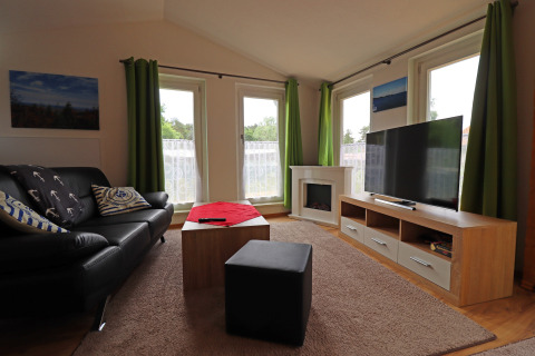 Living room of Räuberkuhle Koserow cabin in Germany with sofa, TV, green curtains and large windows.