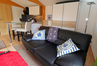 Modern cabin living room at Räuberkuhle Koserow in Germany with black sofa, nautical pillows, and kitchenette.