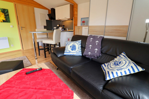 Modern living room at Räuberkuhle Koserow, Germany with a black sofa, nautical pillows, and kitchenette.