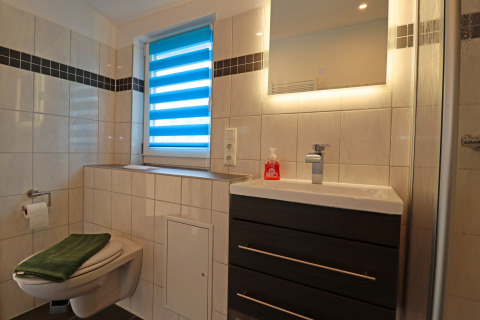 Modern bathroom with wall-mounted toilet, sink cabinet, backlit mirror, and window with blue blinds.