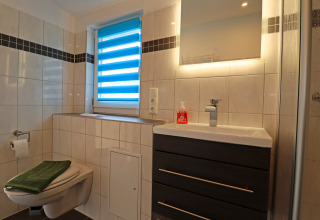 Modern bathroom with wall-mounted toilet, sink cabinet, backlit mirror, and window with blue blinds.