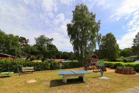 Playground at Räuberkuhle Koserow in Germany featuring ping pong table, swings, and wooden playhouse.