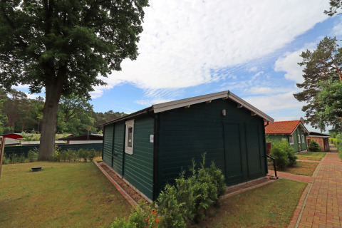 Green wooden cabin at Räuberkuhle Koserow in Germany, surrounded by trees, pathway, and blue sky.