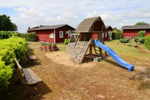 Playground area with a slide, climbing frame, and benches by red cabins in Räuberkuhle Koserow, Germany.
