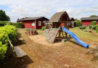 Playground area with a slide, climbing frame, and benches by red cabins in Räuberkuhle Koserow, Germany.