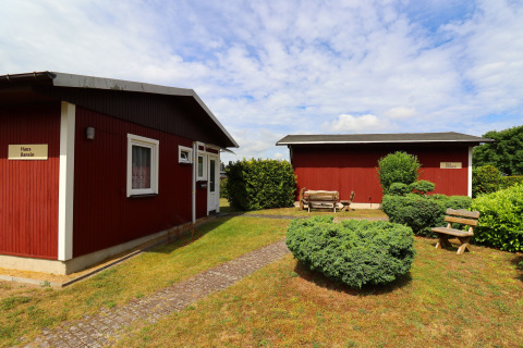 Two red cabins with name signs, benches, and bushes in a garden setting in Räuberkuhle Koserow, Germany.