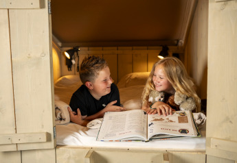 Due bambini leggono un libro e sorridono in un letto accogliente dentro una tenda safari, tra peluche.