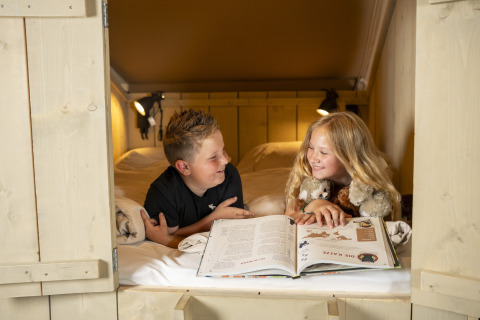 Two kids read a book and share a smile in a cozy bed inside a safari tent, surrounded by stuffed animals.