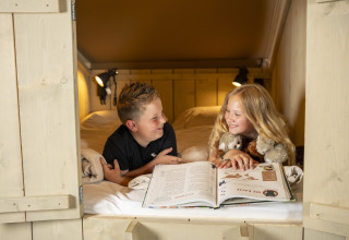 Two kids read a book and share a smile in a cozy bed inside a safari tent, surrounded by stuffed animals.