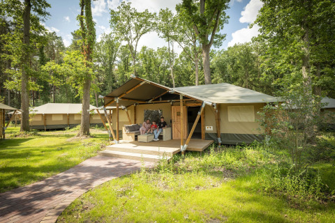 Family relaxing on porch of a safari tent in green forest, other tents visible in the background.