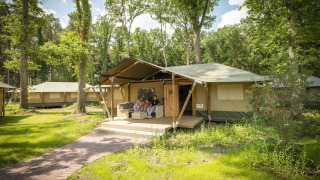 Family relaxing on porch of a safari tent in green forest, other tents visible in the background.