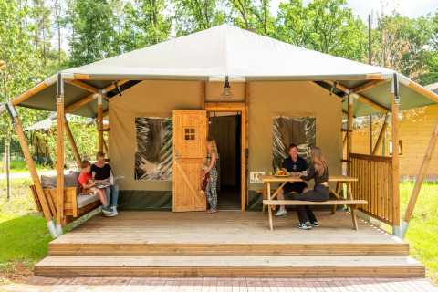 Family relaxing on the porch of a safari tent with sofa and table, surrounded by greenery outdoors.