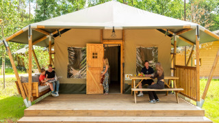 Family relaxing on the porch of a safari tent with sofa and table, surrounded by greenery outdoors.