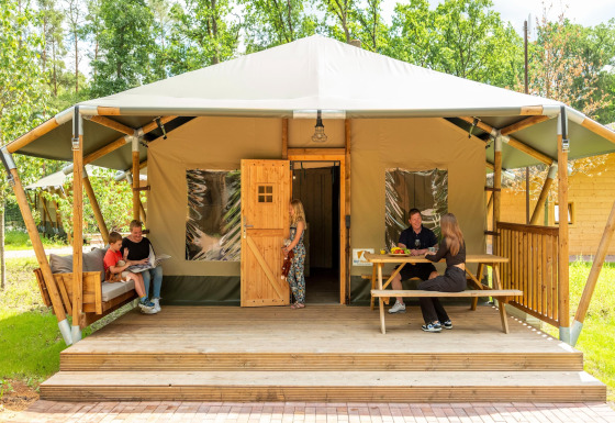 Family relaxing on the porch of a safari tent with sofa and table, surrounded by greenery outdoors.