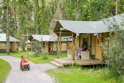 Safari tents in a wooded area, child driving a toy car, and couple relaxing on the porch outside.